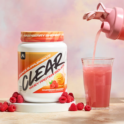 Stance Supplements Clear Whey Protein Isolate being poured into a glass with raspberries on a wooden surface.