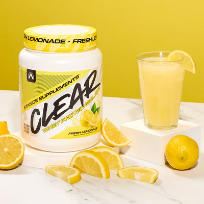 Stance Supplements Clear Whey Protein Powder with a glass of lemonade and lemons on a white surface.